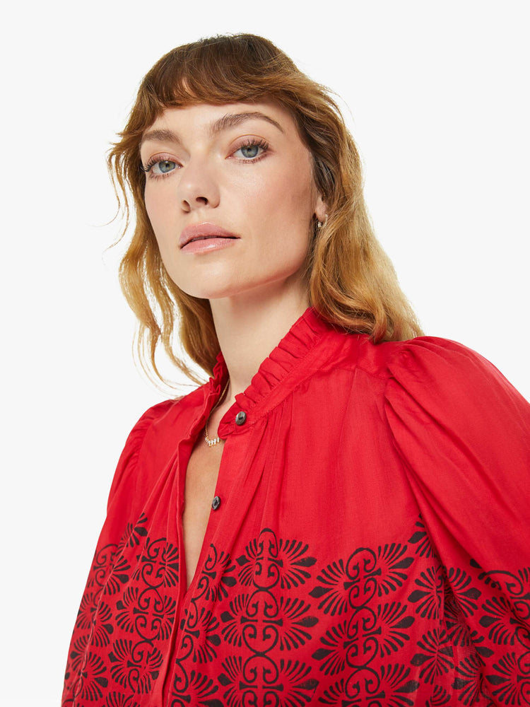 Close up view of a woman blouse in red with black printed details, and features a ruffled collar, puffed shoulders, buttons down the front and a slightly cropped fit.