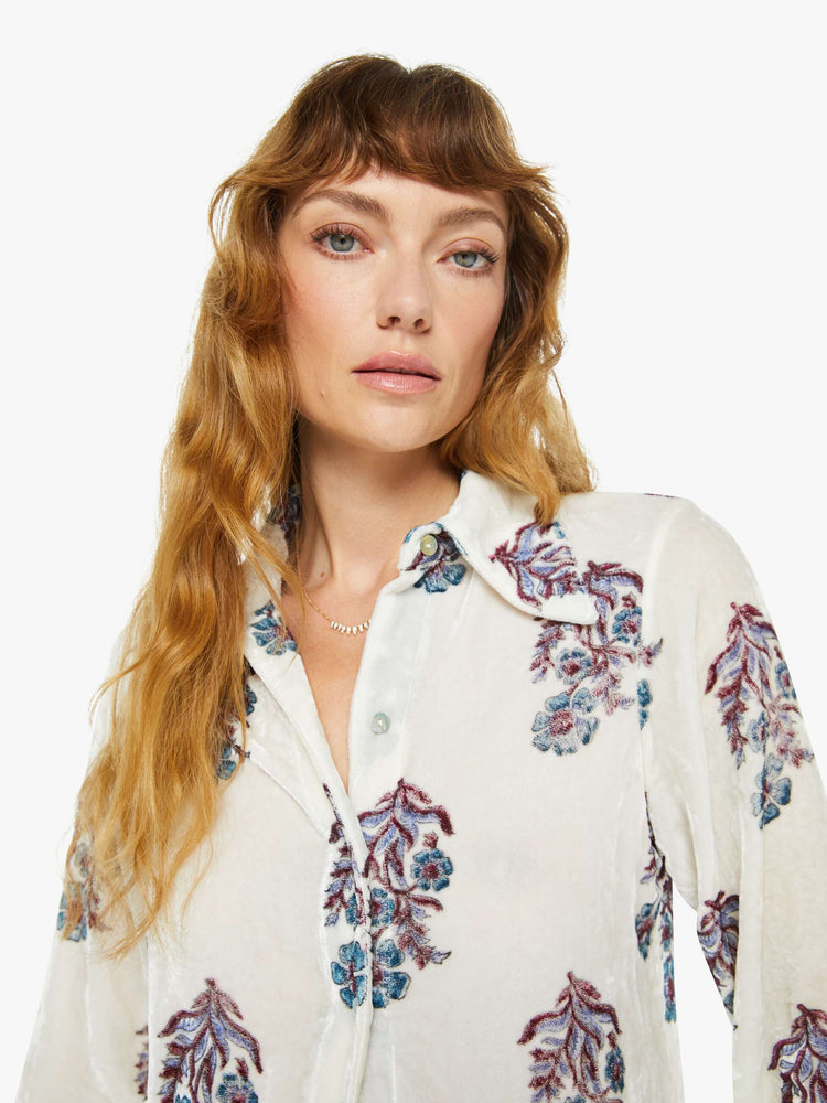 Close up view of a woman blouse in off-white velvet with a cool-toned floral print, and features a collared neck, buttons down the front and long sleeves.