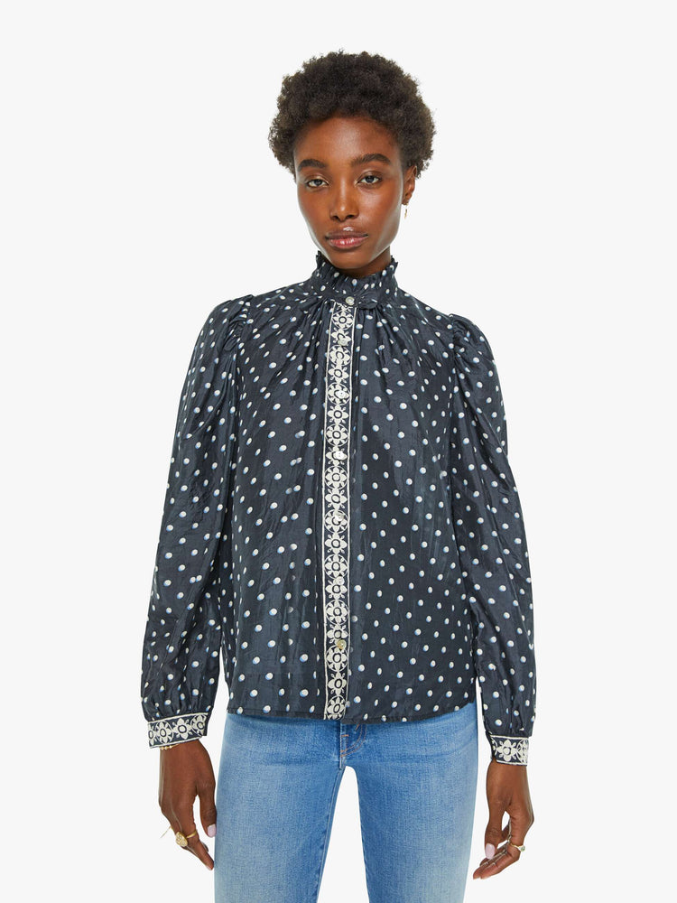 Front view of a blouse in navy with white polka dots and printed trim, and features a ruffled collar, puff shoulders, long balloon sleeves and a loose, flowy fit.