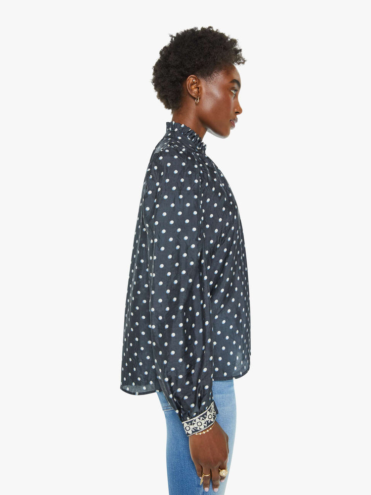 Side view of a blouse in navy with white polka dots and printed trim, and features a ruffled collar, puff shoulders, long balloon sleeves and a loose, flowy fit.