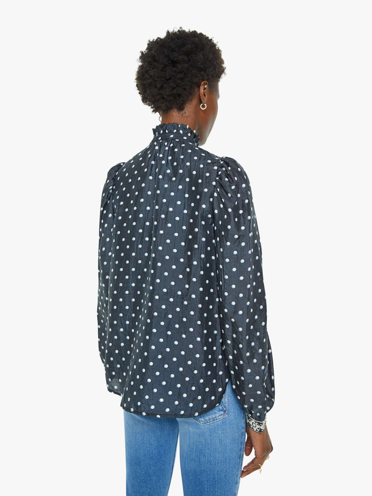 Back view of a blouse in navy with white polka dots and printed trim, and features a ruffled collar, puff shoulders, long balloon sleeves and a loose, flowy fit.