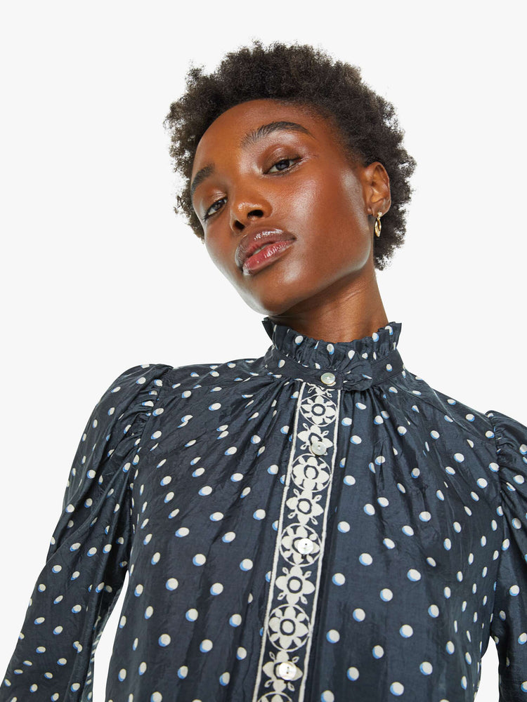 Close up view of a blouse in navy with white polka dots and printed trim, and features a ruffled collar, puff shoulders, long balloon sleeves and a loose, flowy fit.