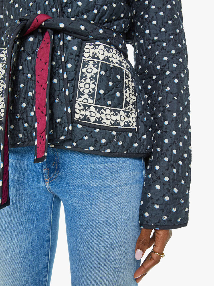 Close up view of a woman jacket  in navy with white polka dots and red trim, and features a V-neck, front patch pockets and a tied waist.