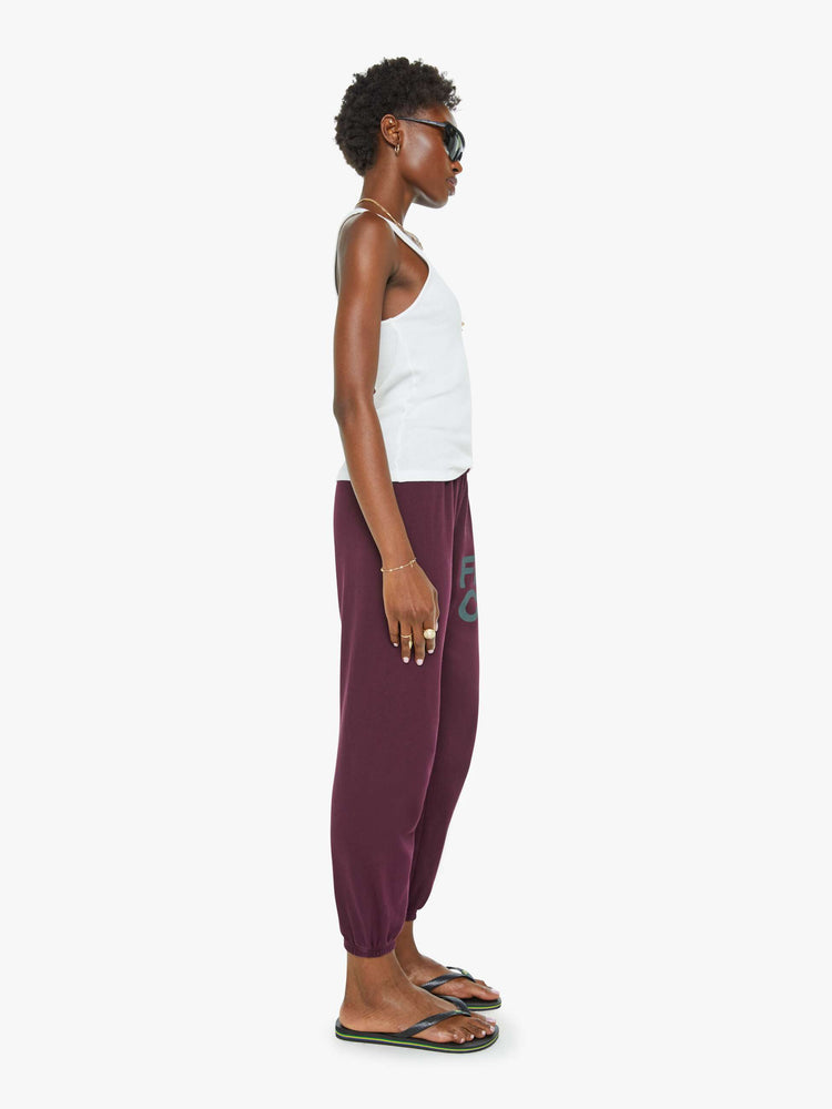 Side view of a woman in an eggplant color sweatpants with a drawstring waistband, elastic hems, a loose, wide leg and the brand's logo in grey at the hip.