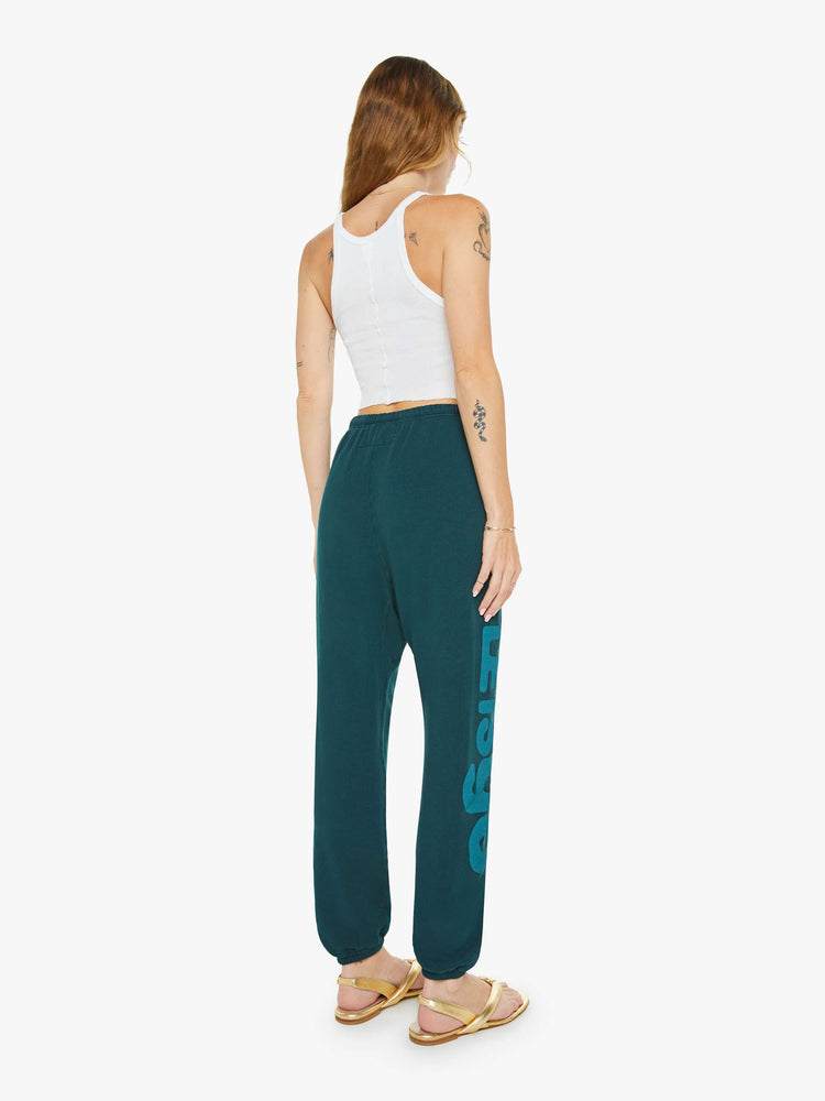 Back view of a woman in sweatpants designed with a drawstring waistband, elastic hems, a loose leg and text on the front and side.