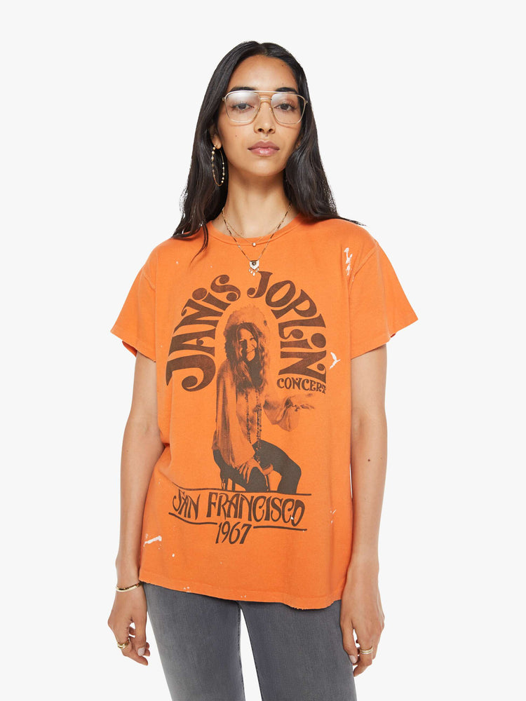 Front view of a woman in a distressed crewneck tee in orange, the tee pays homage to Janis Joplin's 1967 tour with a graphic portrait on the front.