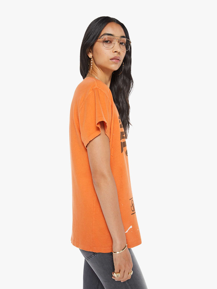 Side view of a woman in a distressed crewneck tee in orange, the tee pays homage to Janis Joplin's 1967 tour with a graphic portrait on the front.