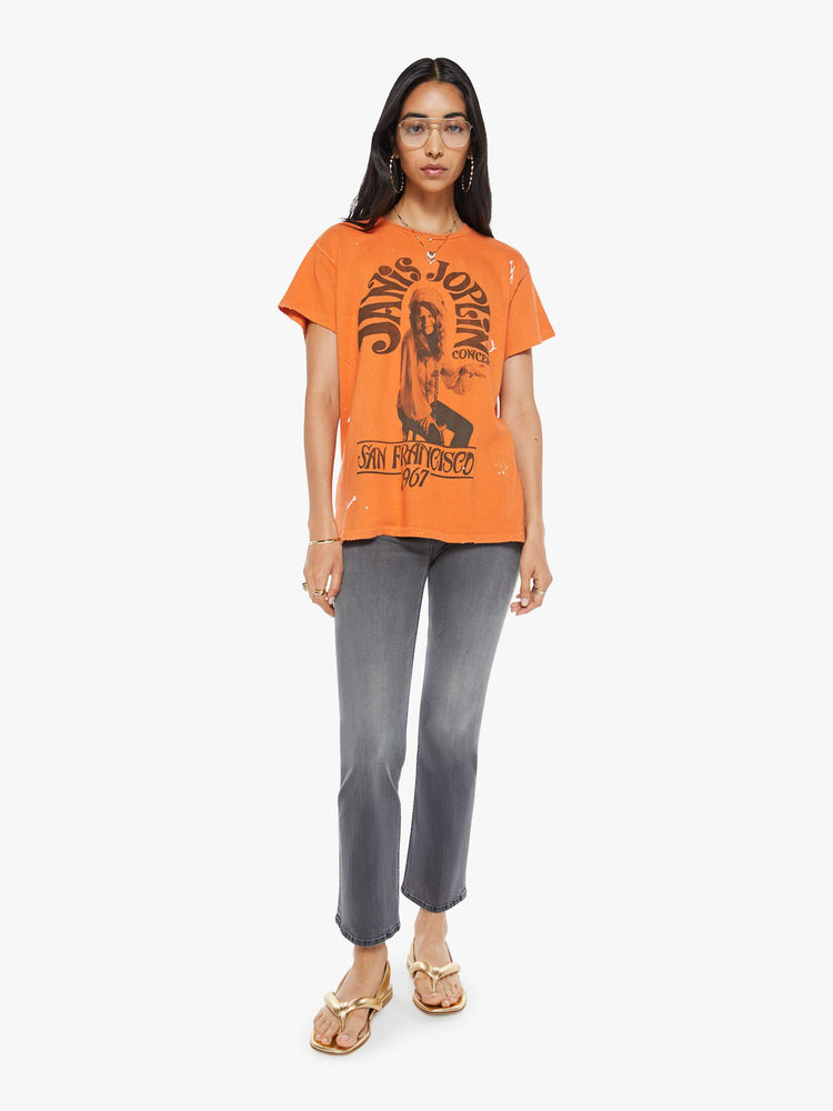 Full body view of a woman in a distressed crewneck tee in orange, the tee pays homage to Janis Joplin's 1967 tour with a graphic portrait on the front.