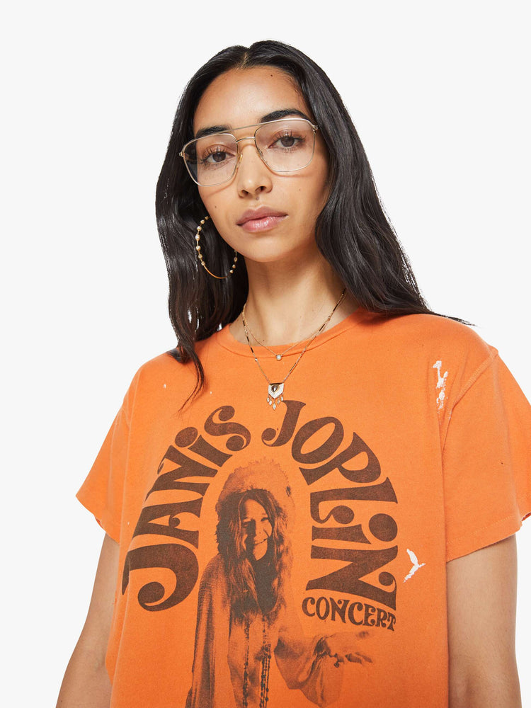 Close up view of a woman in a distressed crewneck tee in orange, the tee pays homage to Janis Joplin's 1967 tour with a graphic portrait on the front.