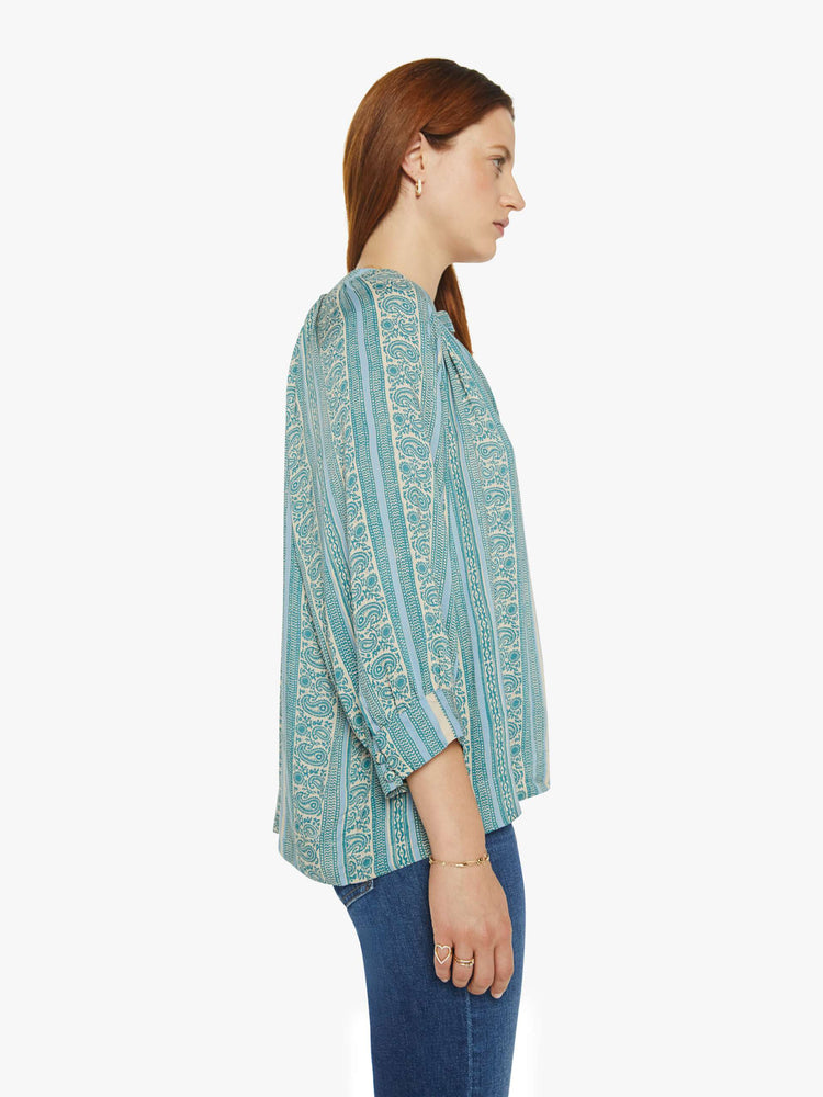 Side view of a woman V-shaped neckline shirt with covered buttons and subtle pleats below the collar emphasizes the loose, flowy feel in a teal stripe pattern.