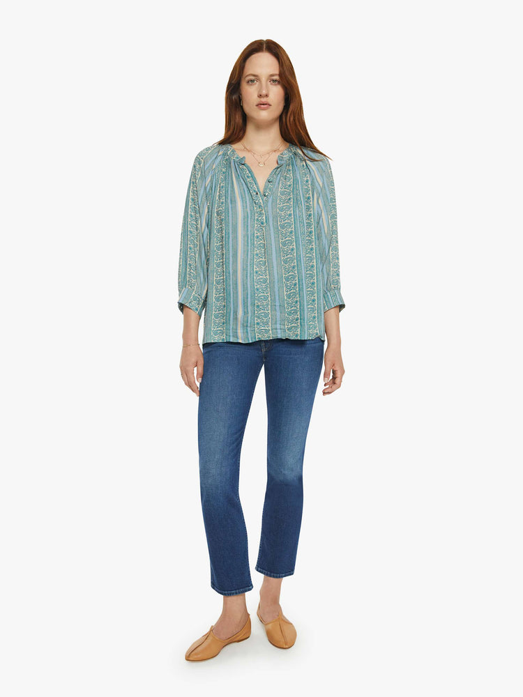 Full body view of a woman V-shaped neckline shirt with covered buttons and subtle pleats below the collar emphasizes the loose, flowy feel in a teal stripe pattern.