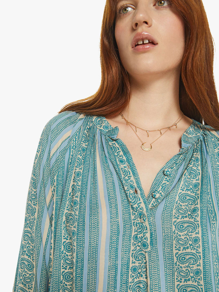 Close up view of a woman V-shaped neckline shirt with covered buttons and subtle pleats below the collar emphasizes the loose, flowy feel in a teal stripe pattern.