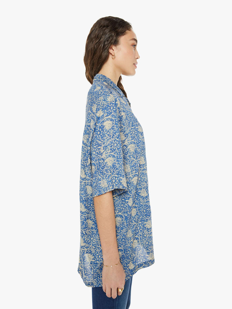 Side view of a woman in a shirt with a designed in a blue floral print, and features a V-neck, drop shoulders, elbow-length sleeves, buttons down the front and a long, thigh-grazing hem.