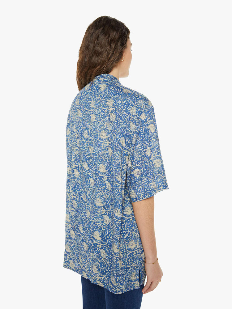 Back view of a woman in a shirt with a designed in a blue floral print, and features a V-neck, drop shoulders, elbow-length sleeves, buttons down the front and a long, thigh-grazing hem.