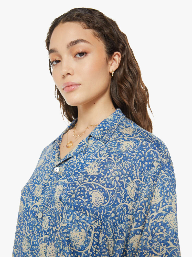 Close up view of a woman in a shirt with a designed in a blue floral print, and features a V-neck, drop shoulders, elbow-length sleeves, buttons down the front and a long, thigh-grazing hem.