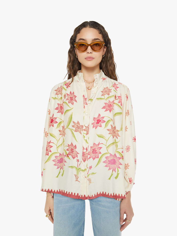 Front view of a woman in cream with a pink floral print long sleeve top has a short stacked collar, a V-neck with buttons down the front, a loose, flowy fit and red trim.