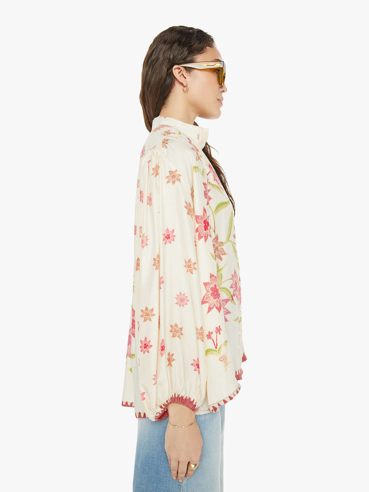 Side view of a woman in cream with a pink floral print long sleeve top has a short stacked collar, a V-neck with buttons down the front, a loose, flowy fit and red trim.