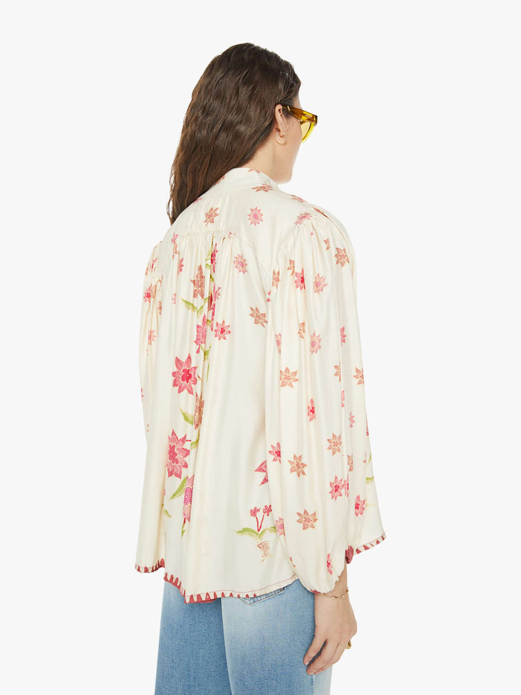 Back view of a woman in cream with a pink floral print long sleeve top has a short stacked collar, a V-neck with buttons down the front, a loose, flowy fit and red trim.