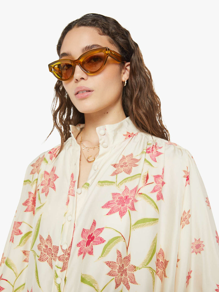 Close up view of a woman in cream with a pink floral print long sleeve top has a short stacked collar, a V-neck with buttons down the front, a loose, flowy fit and red trim.