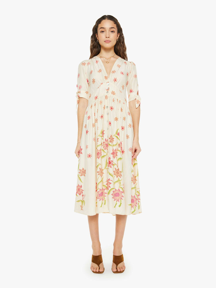 Front view of a woman in a cream with a pink floral print midi dress has a deep V-neck, elbow-length sleeves that tie, buttons down the front and ruffled details throughout.