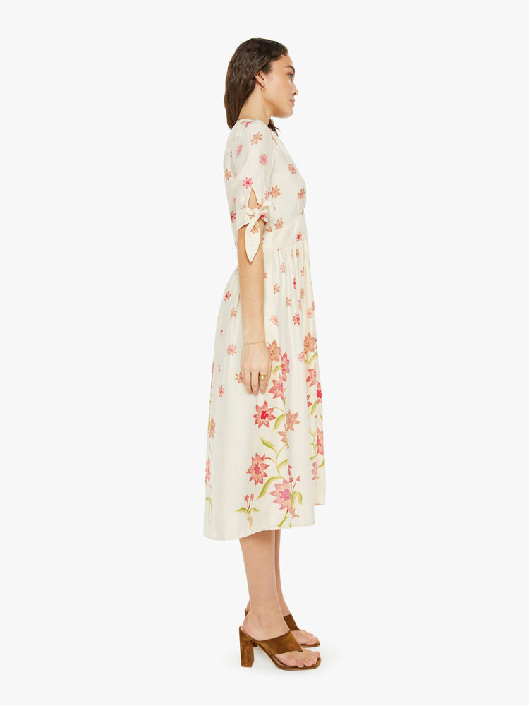 Side view of a woman in a cream with a pink floral print midi dress has a deep V-neck, elbow-length sleeves that tie, buttons down the front and ruffled details throughout.