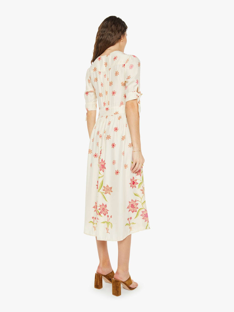 Back view of a woman in a cream with a pink floral print midi dress has a deep V-neck, elbow-length sleeves that tie, buttons down the front and ruffled details throughout.