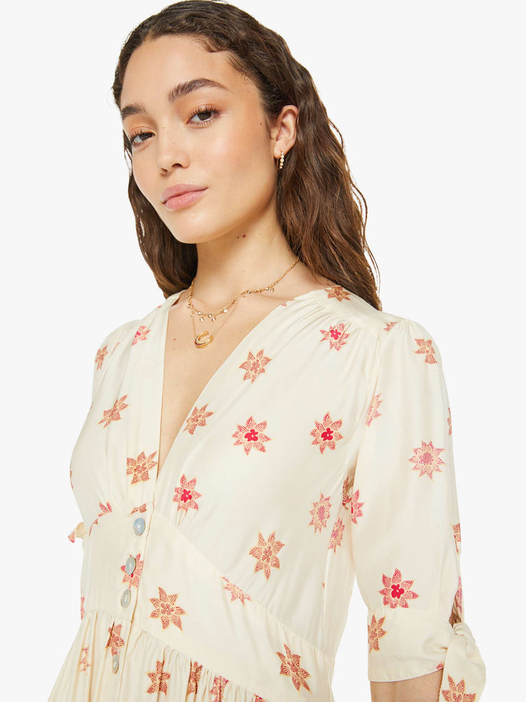 Close up view of a woman in a cream with a pink floral print midi dress has a deep V-neck, elbow-length sleeves that tie, buttons down the front and ruffled details throughout.