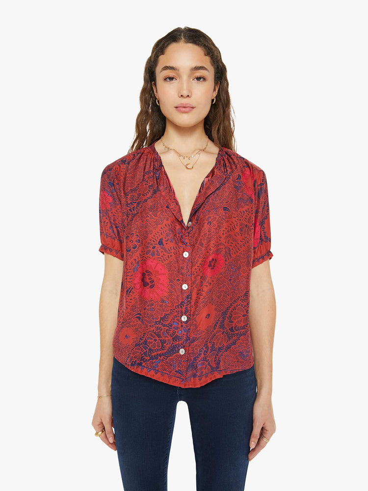 Front view of a woman in a deep red with blue floral print top with a deep V-neck, buttons down the front, short balloon sleeves and ruffles throughout for a loose, flowy fit.