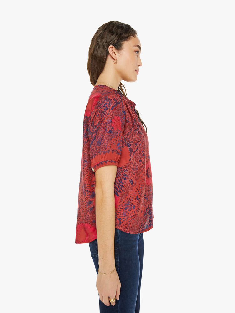 Side view of a woman in a deep red with blue floral print top with a deep V-neck, buttons down the front, short balloon sleeves and ruffles throughout for a loose, flowy fit.