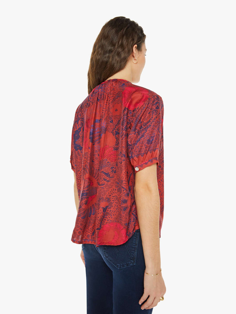 Back view of a woman in a deep red with blue floral print top with a deep V-neck, buttons down the front, short balloon sleeves and ruffles throughout for a loose, flowy fit.