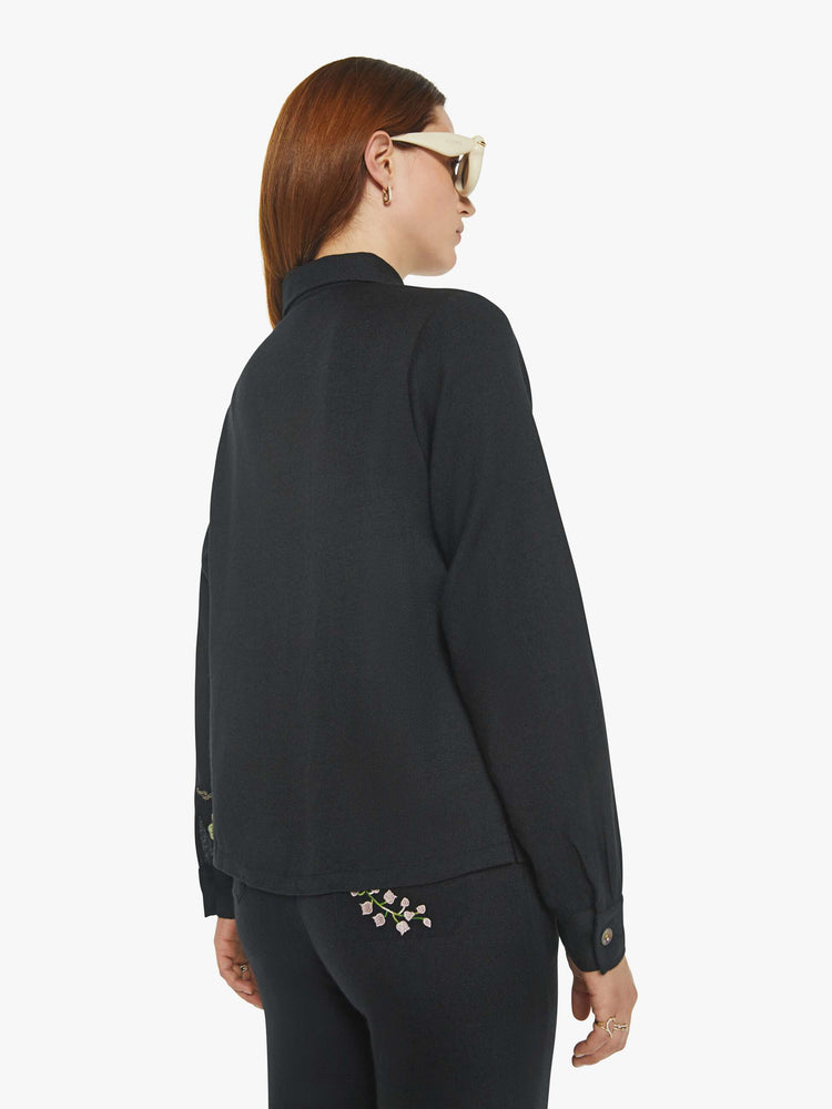 Back view of a woman in a black shirt with lily of the valley plants along the collar and chest, and features extra-long sleeves and a loose fit.