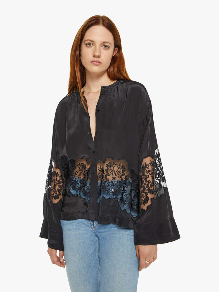 Front view of a woman in a black with lace details at the waist and sleeves blouse with long loose sleeves, a flowy fit and buttons down the front.