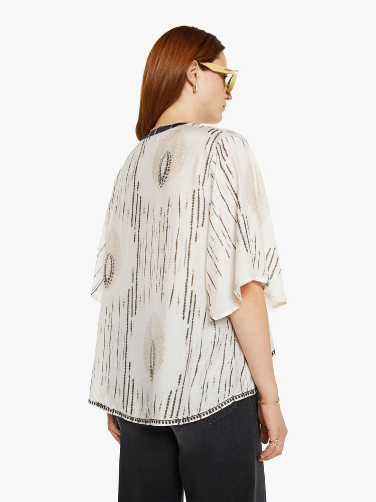 Back view of a woman in cream with delicate black print blouse with a  deep V-neck, boxy elbow-length sleeves and a loose fit. 