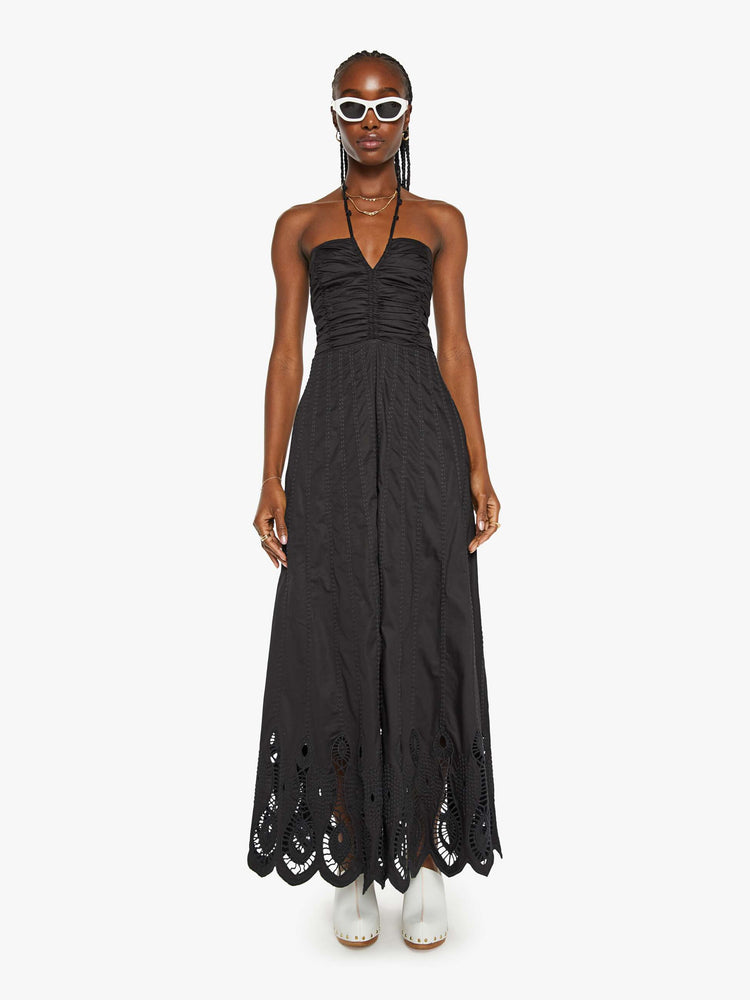 Front view of a woman in a black maxi dress a halter neck, gathered bodice and a floor-length hem with eyelet details.