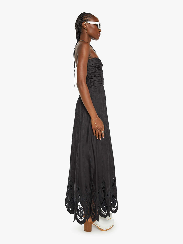 Side view of a woman in a black maxi dress a halter neck, gathered bodice and a floor-length hem with eyelet details.