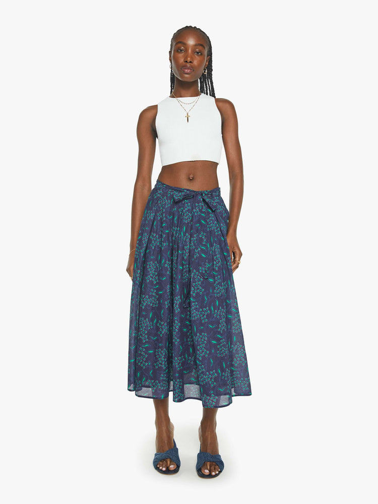 Front view of a woman in a midi-skirt with blue green floral print with a calf-length hem and pleats at the waist.