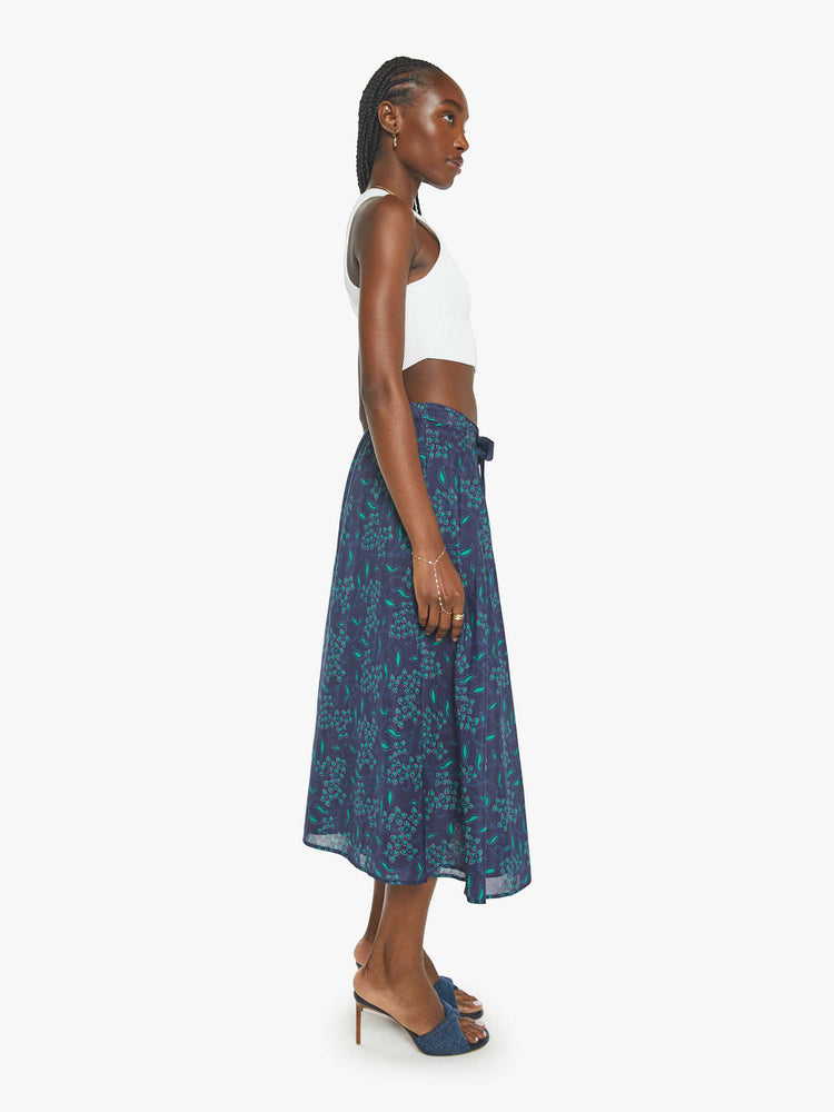 Side view of a woman in a midi-skirt with blue green floral print with a calf-length hem and pleats at the waist.