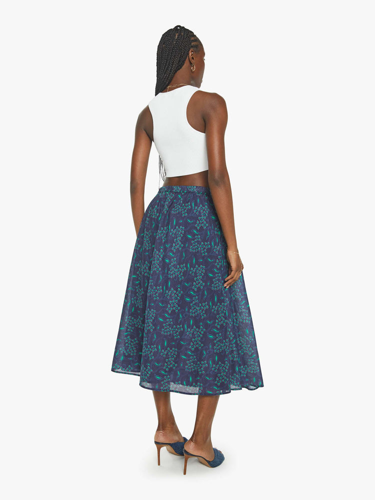 Back view of a woman in a midi-skirt with blue green floral print with a calf-length hem and pleats at the waist.