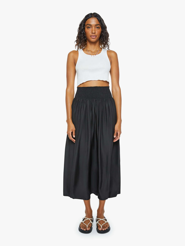 Front view of a woman wearing a black skirt featuring a wide elastic waistband and a flowy fit, paired with a cropped white tank top.