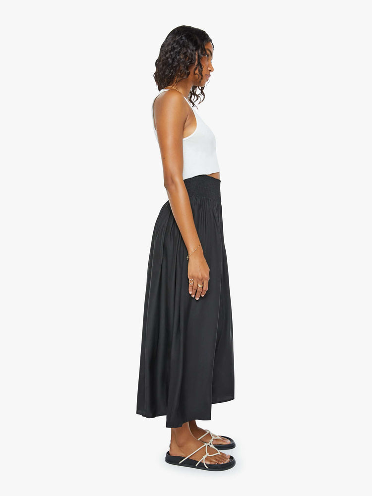 Side view of a woman wearing a black skirt featuring a wide elastic waistband and a flowy fit, paired with a cropped white tank top.