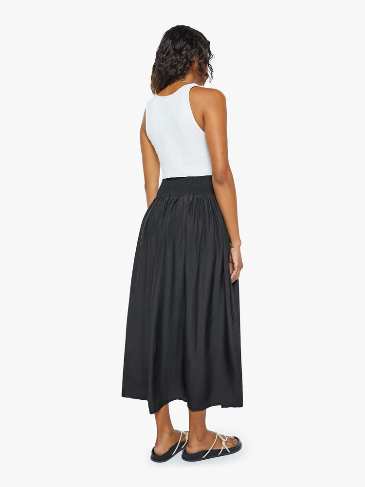 Back view of a woman wearing a black skirt featuring a wide elastic waistband and a flowy fit, paired with a cropped white tank top.