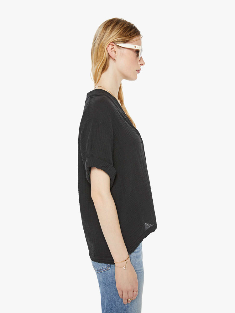 Side view of a woman wearing a black top featuring a deep v-neck, rolled sleeves, and a boxy fit.