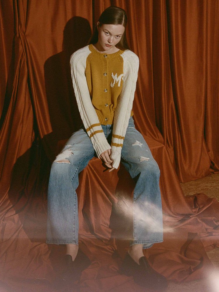 An editorial image of a woman sitting, wearing distressed jeans and a collegiate inspired cardigan.