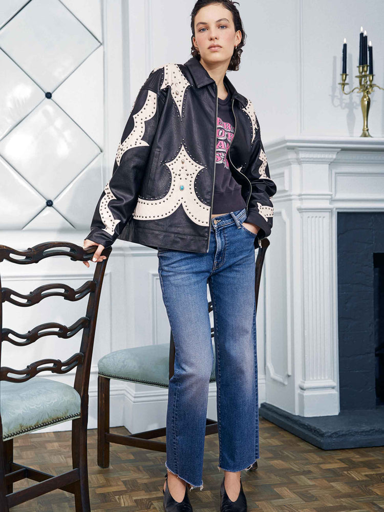 An editorial image of a woman leaning on a chair, wearing a black and white leather jacket with studs, paired with a medium blue wash jean with a frayed hem.