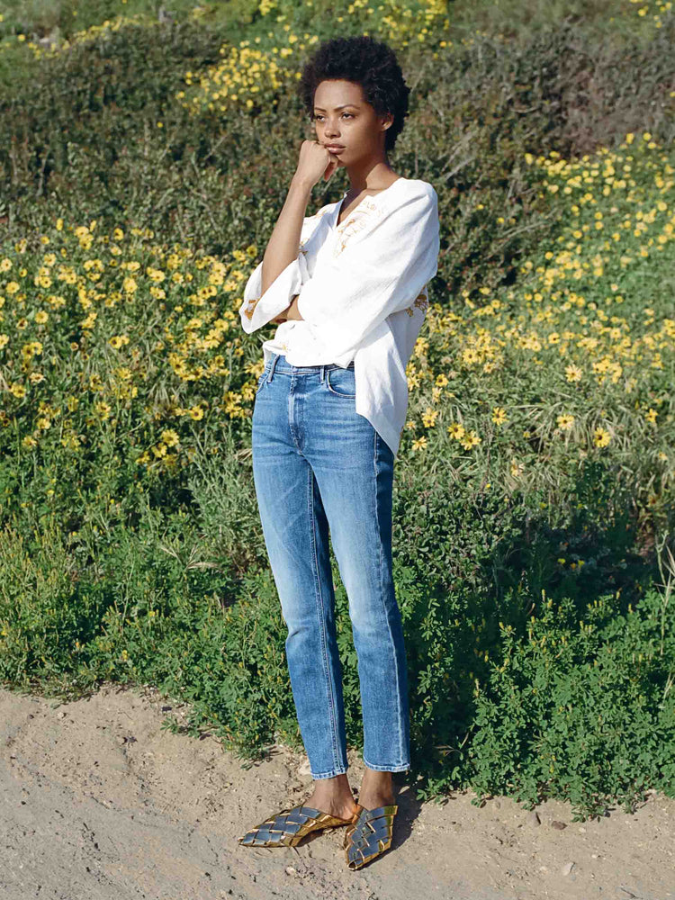 Editorial image of a woman standing in front of a hill of flowers, wearing a white top and medium blue wash jeans.