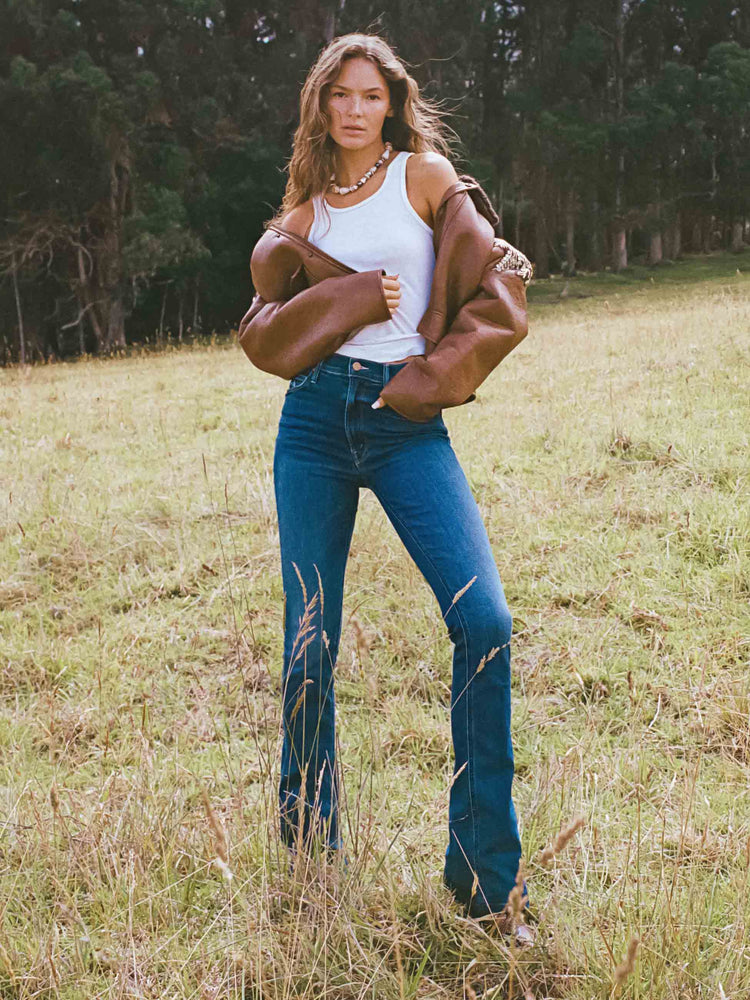 A woman standing in front of a forest, wearing a dark wash flare jean, a white tank, and a frown jacket.